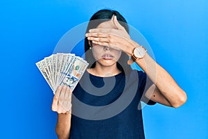 Beautiful young woman holding dollars covering eyes with hand, looking serious and sad