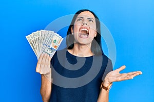 Beautiful young woman holding dollars crazy and mad shouting and yelling with aggressive expression and arms raised