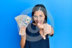 Beautiful young woman holding dollars pointing to you and the camera with fingers, smiling positive and cheerful
