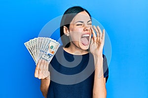 Beautiful young woman holding dollars shouting and screaming loud to side with hand on mouth