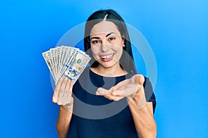 Beautiful young woman holding dollars smiling cheerful offering palm hand giving assistance and acceptance