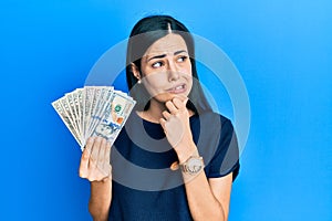 Beautiful young woman holding dollars thinking worried about a question, concerned and nervous with hand on chin