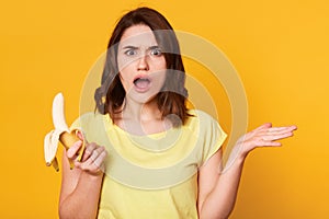 Beautiful young woman holding fresh banana, takes hands to side, has uncomprehending facial expresion, standing with open mouth