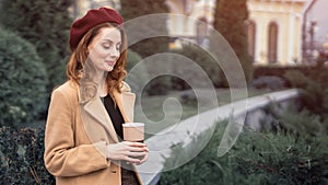 Beautiful young woman holding a mug with coffee standing outdoors. Portrait of stylish young woman wearing autumn coat