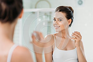 beautiful young woman holding perfume bottle and looking at mirror