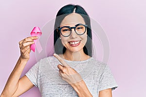 Beautiful young woman holding pink cancer ribbon scared and amazed with open mouth for surprise, disbelief face