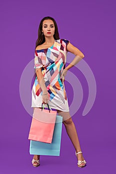 Beautiful young woman holding shopping bags and looking at the camera while standing against purple background.