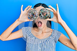 Beautiful young woman holding tasty colorful doughnuts on eyes afraid and shocked with surprise and amazed expression, fear and