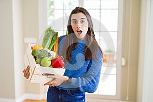 Beautiful young woman holding wooden box full of healthy groceries scared in shock with a surprise face, afraid and excited with
