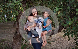 A beautiful young woman hugs her laughing daughter and holds her son `s hand. Family on a walk in the apple garden