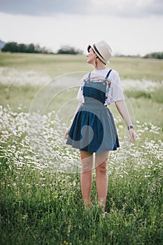 Beautiful young woman in a jeans dress and straw hat posing in a camomile field