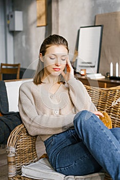A beautiful young woman in jeans and a jumper sits on a wicker chair by the fireplace at home and dreams