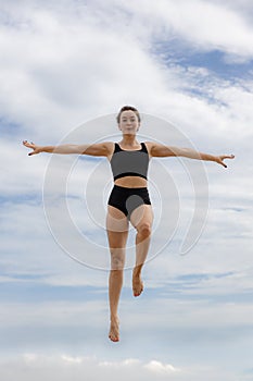 Beautiful young woman jumping over cloudy blue sky. Caucasian woman wearing black sportswear. Fitness, wellness concept. Outdoor