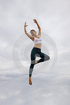 Beautiful young woman jumping over cloudy sky. Caucasian woman wearing sportswear. Fitness, wellness concept. Outdoor activity.
