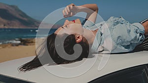 Beautiful young woman laying on car roof carefree. Closeup of car on magnificent beach. Girl enjoying life touching her