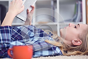 Beautiful young woman laying on the floor at home, reading a book, living room interior. Girl reading a book on the