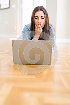 Beautiful young woman laying on the floor using laptop cover mouth with hand shocked with shame for mistake, expression of fear,