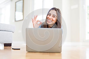 Beautiful young woman laying on the floor using laptop doing ok sign with fingers, excellent symbol