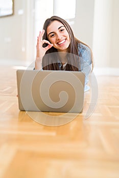 Beautiful young woman laying on the floor using laptop doing ok sign with fingers, excellent symbol