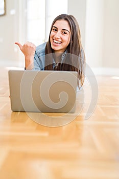 Beautiful young woman laying on the floor using laptop pointing and showing with thumb up to the side with happy face smiling