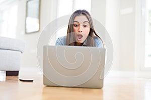 Beautiful young woman laying on the floor using laptop scared in shock with a surprise face, afraid and excited with fear