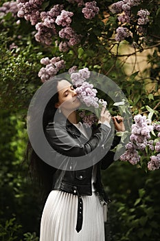 Beautiful young woman in leather jacket enjoying the smell of the bloomy lilac tree in springtime