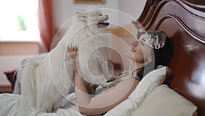 A beautiful young woman lies on the bed and holds a white Pomeranian Spitz in her hands. The girl smiles and looks at