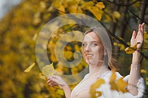 Beautiful young woman with light brown hair in a white sweater on a background of foliage in an autumn park