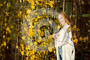 Beautiful young woman with light brown hair in a white sweater on a background of foliage in an autumn park.