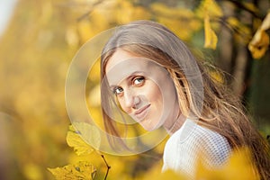 Beautiful young woman with light brown hair in a white sweater on a background of foliage in an autumn park.