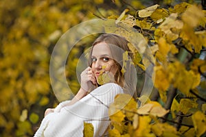 Beautiful young woman with light brown hair in a white sweater on a background of foliage in an autumn park.