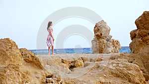 A beautiful young woman in a light dress, fluttering in the wind, is walking along the rocky seashore. against the