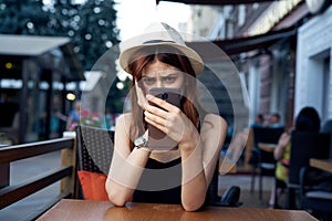 Beautiful young woman in a light hat at a table in the summer in a cafe on the terrace holding a phone