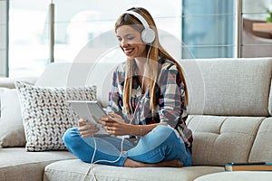 Beautiful young woman listening to music with headphones while using her digital tablet sitting on couch at home