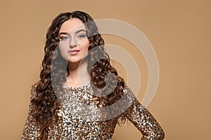 Beautiful young woman with long curly brown hair in golden sequin dress on beige background