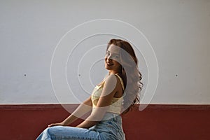 Beautiful young woman with long curly hair against a white and red background. The woman is sitting on a stool in the street. The