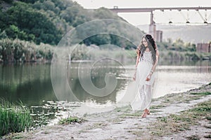 Beautiful young woman with long curly hair dressed in boho style dress posing near lake