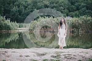 Beautiful young woman with long curly hair dressed in boho style dress posing near lake