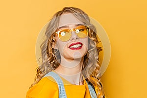 Beautiful young woman with long curly hair wearing sunglasses smiling on yellow background