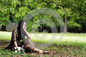 Beautiful young woman in a long dark dress sitting on grass under a tree