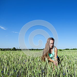 Beautiful young woman with long healthy brown hair, crouching in a field of flowering grain