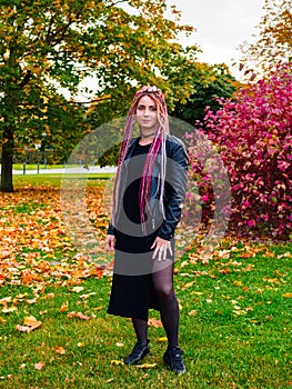 Beautiful young woman with long pink dreadlocks in black dress, biker jacket and sneakers posing in the park on the grass in