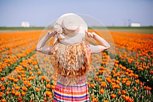 Beautiful young woman with long red hair wearing a striped dress and straw hat standing by the back on colorful tulip field