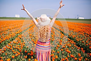 Beautiful young woman with long red hair wearing a striped dress and straw hat standing by the back on colorful tulip field