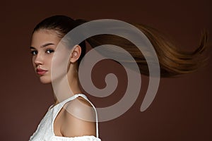 Beautiful young woman with long straight hair and makeup. The hairstyle is gathered in a ponytail. Brown background