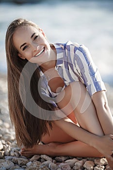 A beautiful young woman with long straight hair sits alone on stones by the sea.