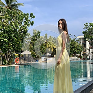 A beautiful young woman in a long yellow dress stands in front of a green pool and palm trees.