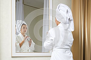 Beautiful young woman look in mirror applying cream, pretty lady wrap towel on head put facial moisturizer lifting