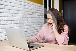 Beautiful young woman looking at laptop and expressing shock sitting at table in room at home