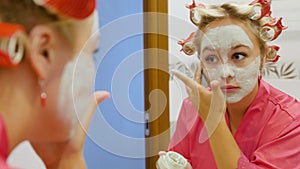 Beautiful young woman looks in the mirror and applies a cosmetic face mask. A pretty blonde in curlers on her head is